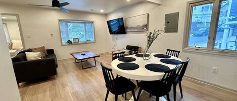 A modern living room with a round dining table, black chairs, a ceiling fan, a TV mounted on the wall, and a window with blinds.