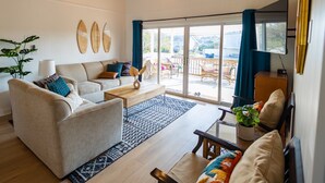 Un salón amplio con techo de madera, un gran ventanal corredero que da a un patio exterior, y una zona de estar acogedora con sofá y sillones.