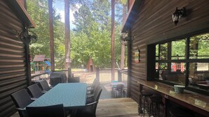 A covered outdoor seating area with a table and chairs, a bar, and a view of the surrounding trees.