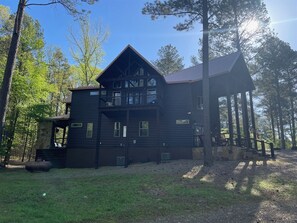 Een ruime, donkere houten chalet met meerdere ramen en een balkon, omgeven door bomen en een heldere lucht.