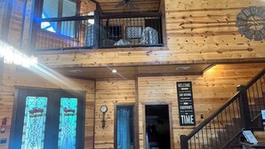 A wooden interior with a staircase, a dining area with a table and chairs, and a sign on the wall that reads "WELCOME IN THE WOODS TAKE NOTHING LEAVE NOTHING TIME".
