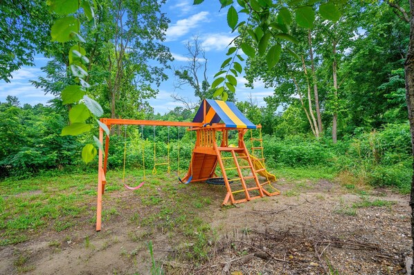 Une structure de jeu en bois, dotée d’une échelle et d’un toboggan, entourée d’arbres et de verdure.