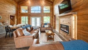 Un intérieur de chalet chaleureux avec une cheminée, des canapés en cuir, un coin repas et un téléviseur fixé au mur.