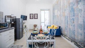 A modern kitchen with a dining table, chairs, and a chandelier. There is a blue sofa with yellow cushions in the living area. - Elizabeth House (Siracusa)