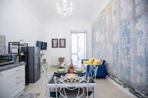 A modern kitchen with a dining table, chairs, and a chandelier. There is a blue sofa with yellow cushions in the living area.