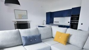 Una sala de estar moderna con un sofá modular gris, una mesa de centro negra y una cocina con armarios azules.
