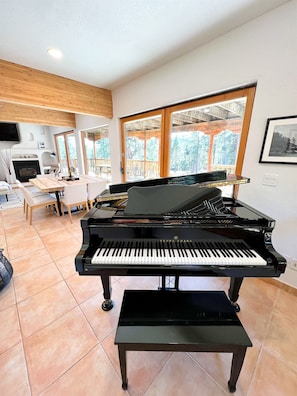 A grand piano in a spacious living area with a fireplace, large windows, and a wooden ceiling.