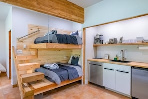 A bunk bed with a wooden headboard, a small wooden bench, a kitchen area with a sink, a coffee maker, and a dishwasher.