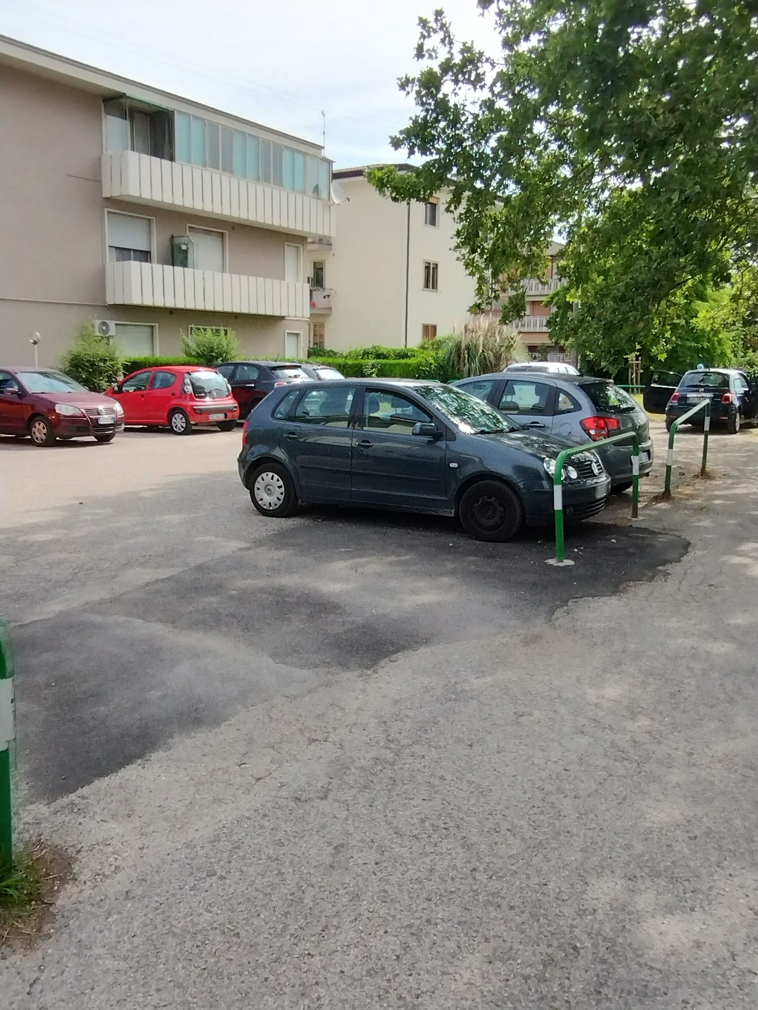 A parking lot with several cars parked, a building with a balcony, and a tree in the background.