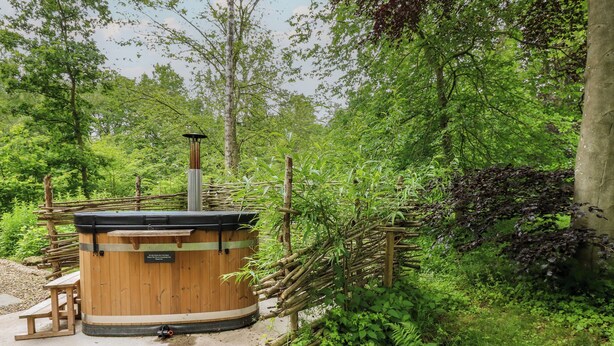 A wooden hot tub with a label, surrounded by a natural setting with trees and a wooden fence. - Bracken Fern (Richmond)
