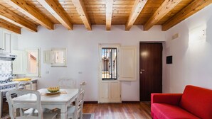 A room with a wooden ceiling, a dining table with chairs, a red sofa, and a kitchen area with a stove and cabinets.