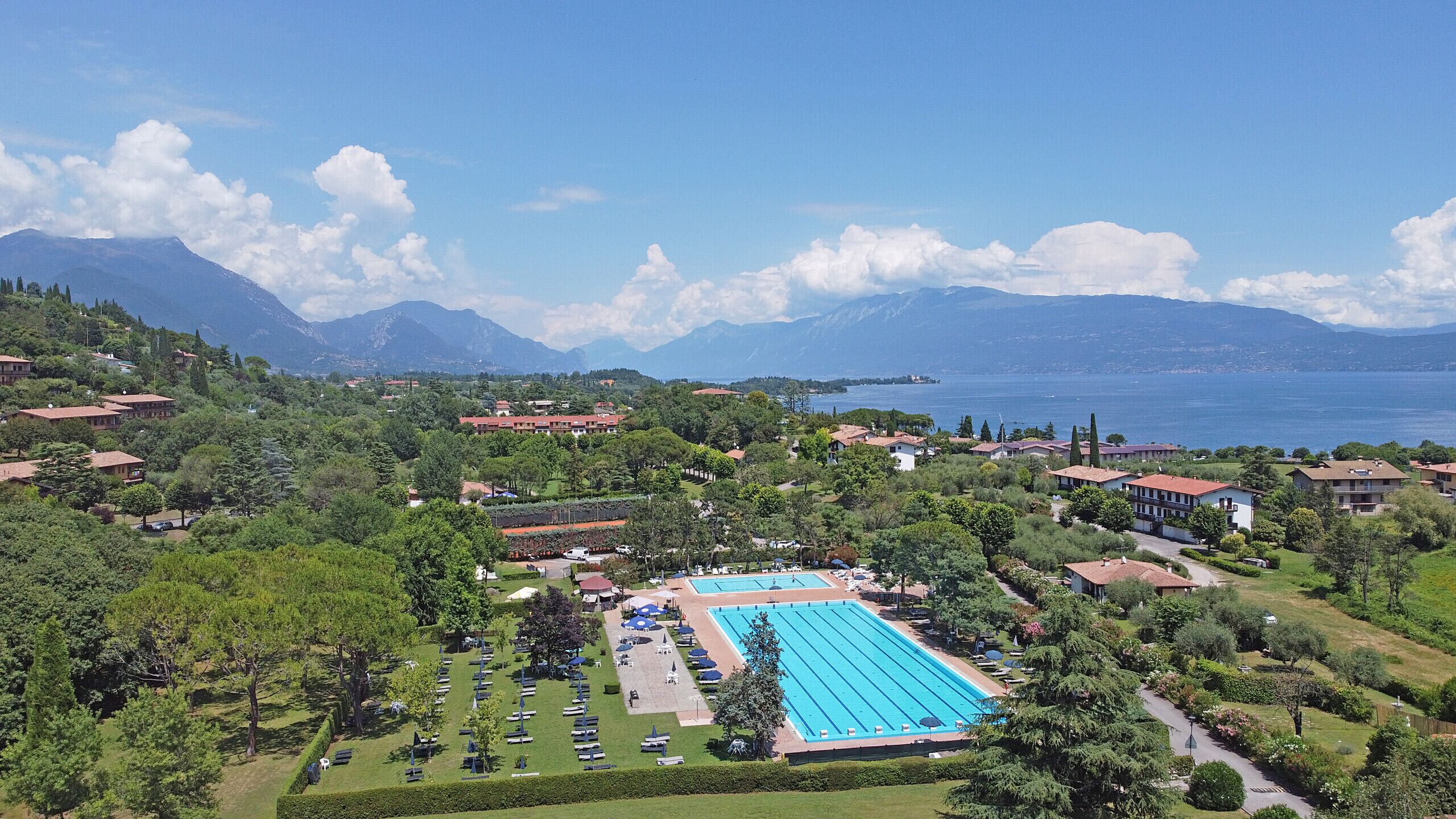 A resort with a large swimming pool, lush greenery, and a view of a lake and mountains.