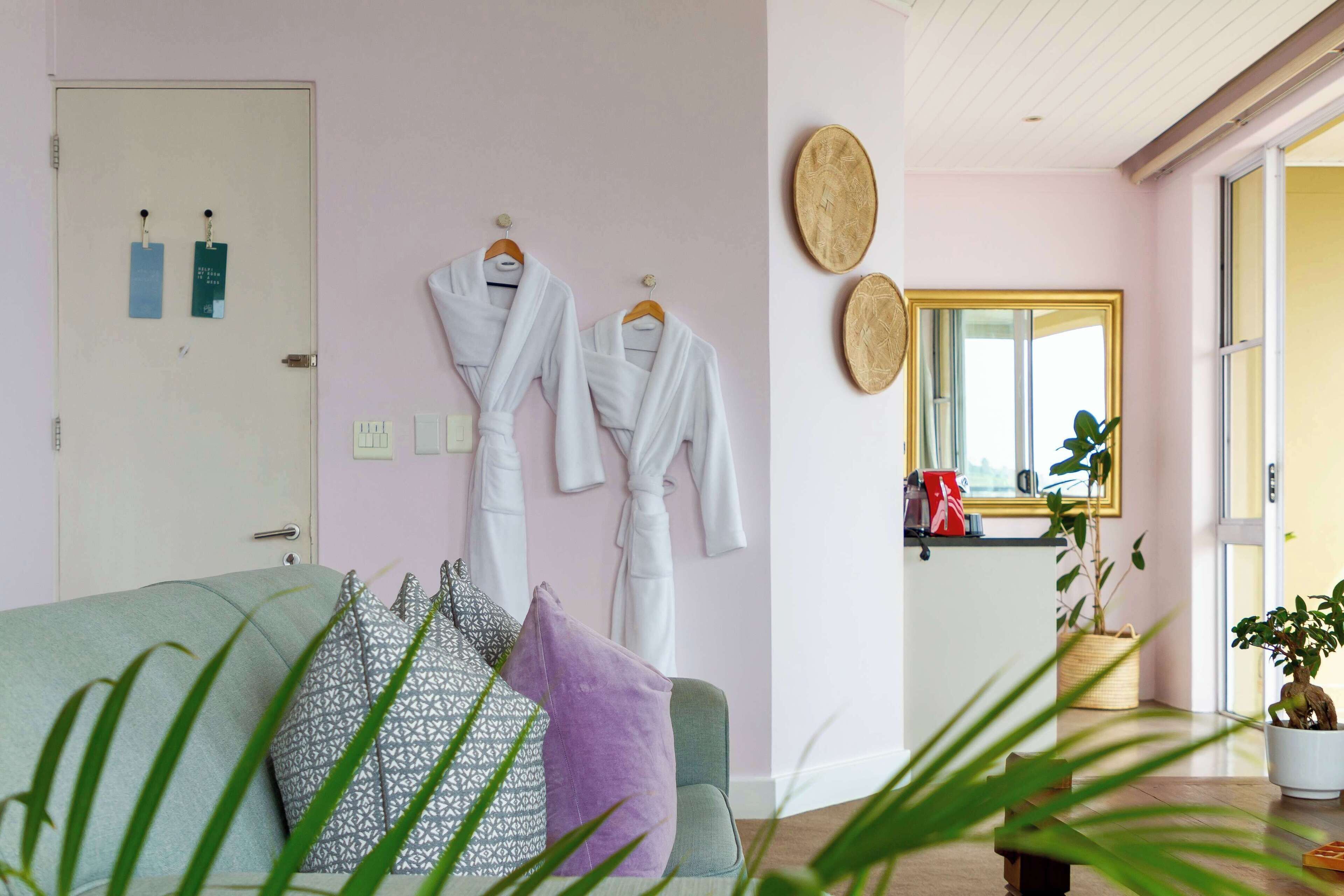A room with a green sofa, a purple pillow, and a white robe hanging on the wall. There is a door with two ID badges, a mirror, and a window with a view of the outside.