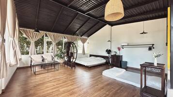 A spacious room with a large bed, a seating area with a table and chairs, a hanging light fixture, and a view of greenery outside.