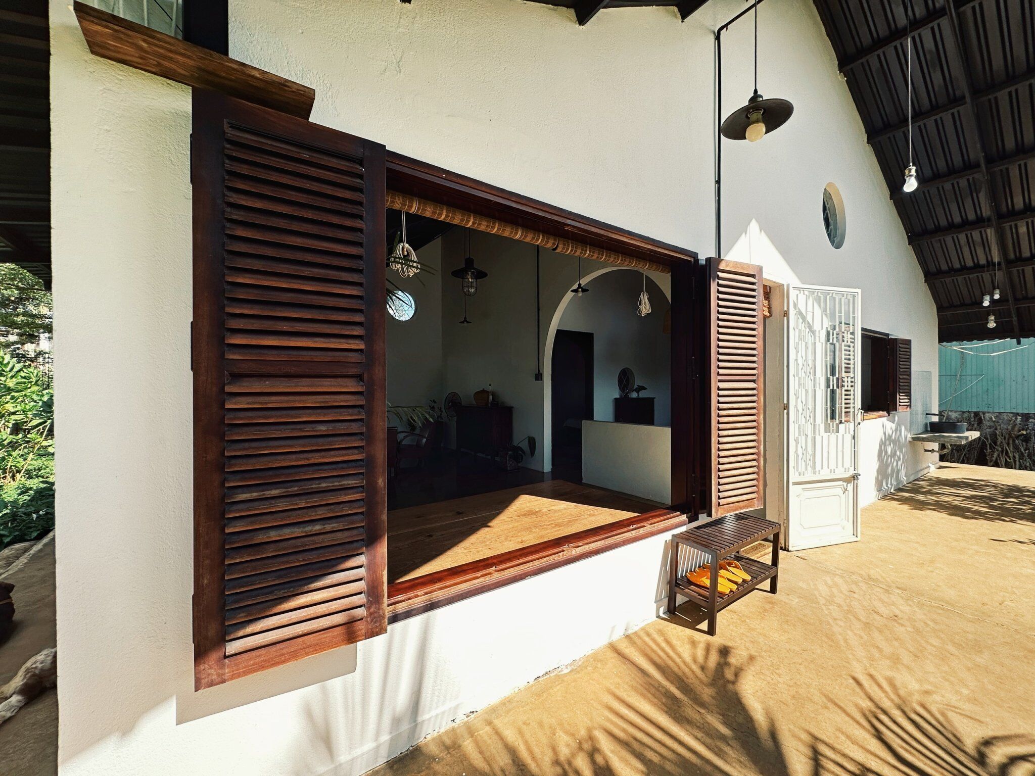 A room with a large window, wooden shutters, a hanging light fixture, and a ceiling fan.