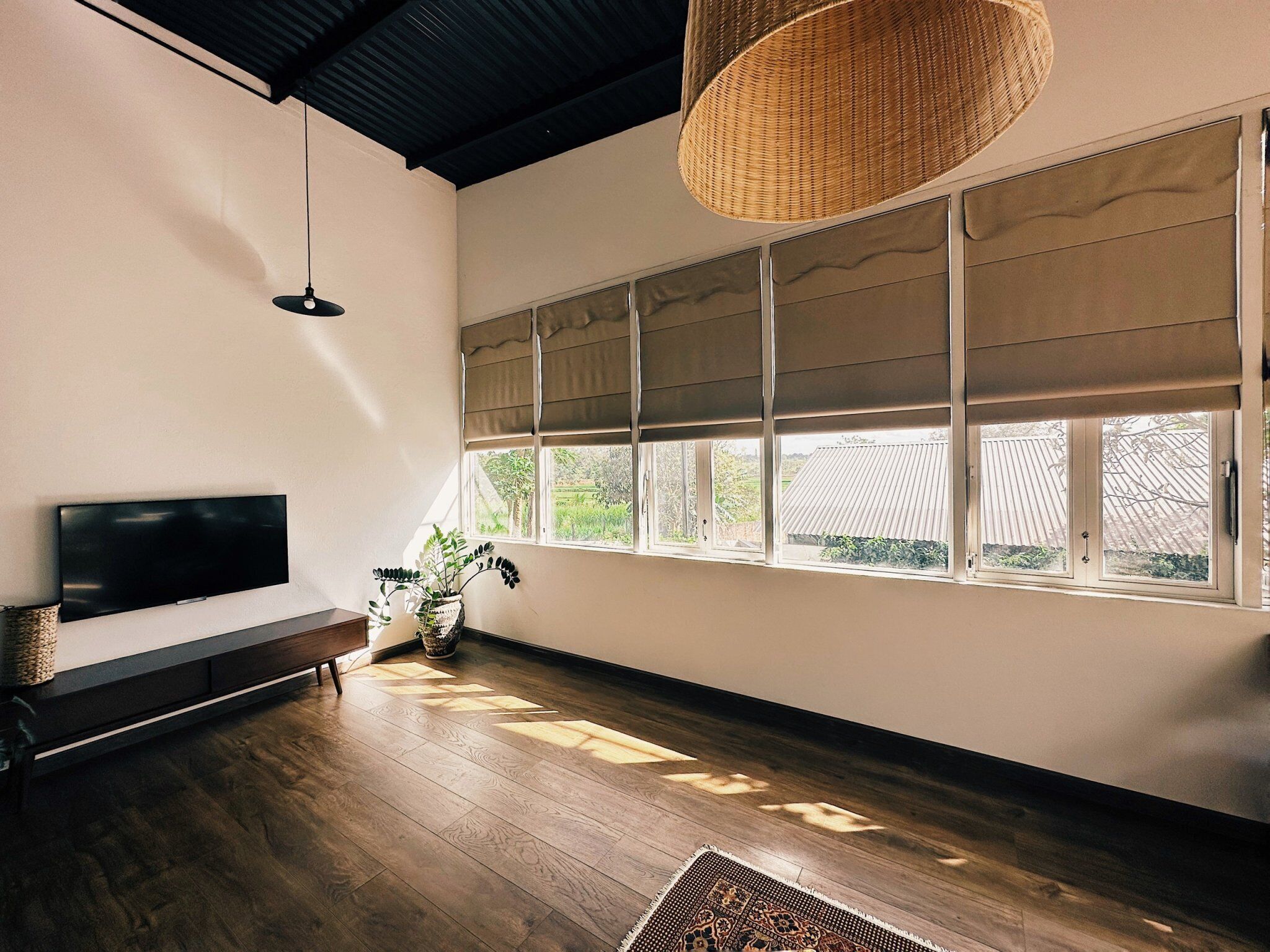 A modern living room with a flat-screen TV, a large window with blinds, a woven light fixture, and a patterned rug.