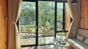 A room with a large sliding glass door leading to a garden, a sofa, a small table with a tray, and a lamp. - An Nhien Farmstay & Resort (Pleiku)