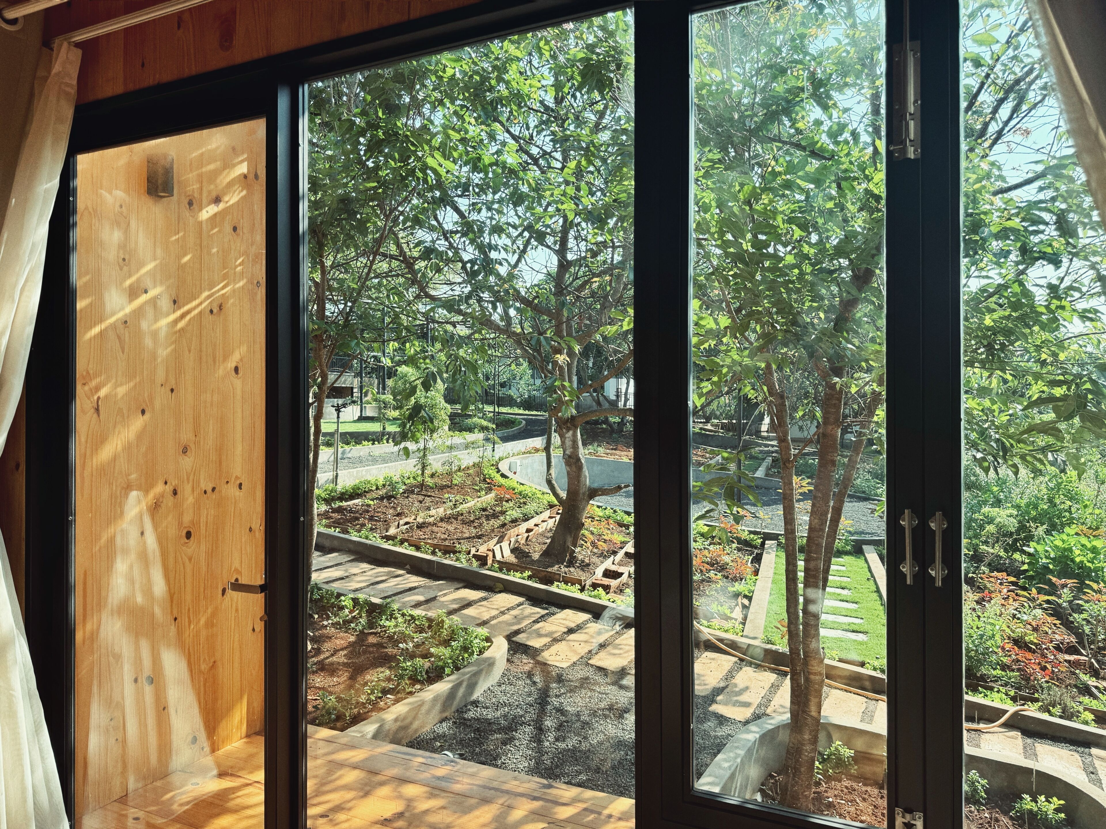Vista desde una habitación con un ventanal grande que da a un jardín con árboles y un camino pavimentado.