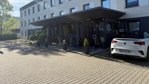Un bâtiment moderne doté d’une entrée couverte, une voiture blanche garée devant et une allée pavée.