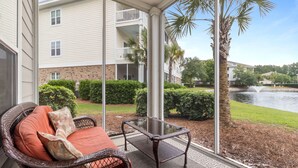 A wicker sofa with cushions, a glass-top table, and a view of a building and water feature. - Willow Bend #1411 Nice Condo! (North Myrtle Beach)