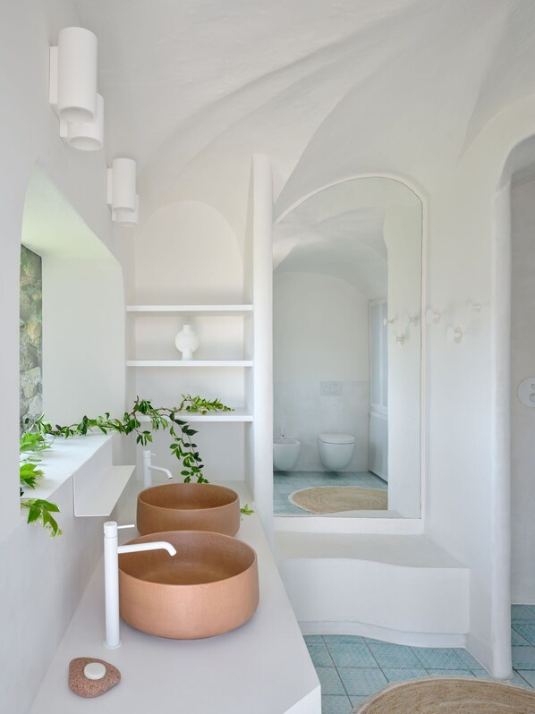 Un baño moderno con una paleta de colores blanco, que presenta un lavamanos de cobre grande, un lavamanos pedestal blanco y un espejo sobre los lavamanos. Hay estantes con plantas y una vista al área de la ducha.