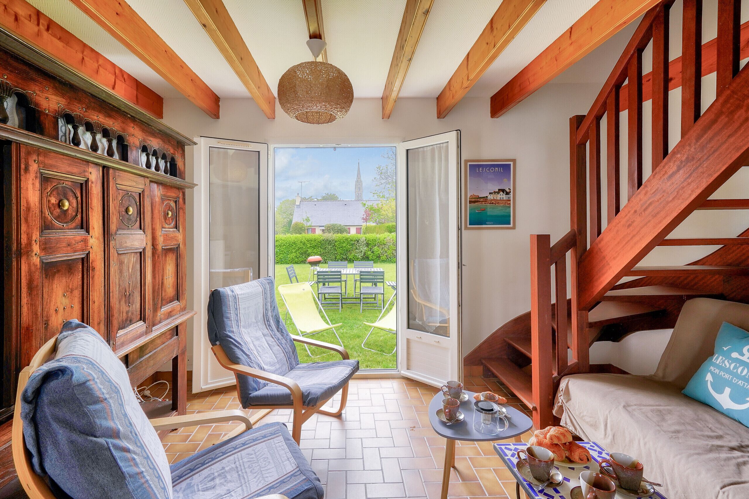 A cozy room with a wooden ceiling, a large window, a sofa, a chair, a small table with cups and a teddy bear, and a cabinet with decorative items.