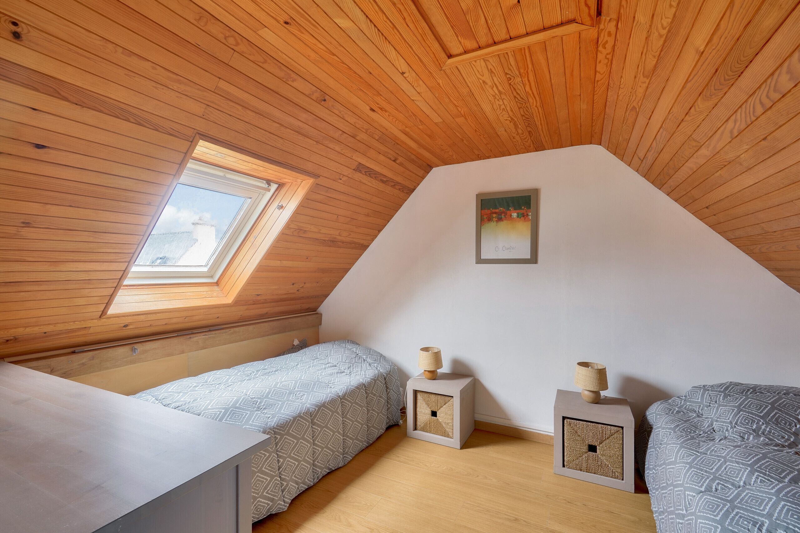 Een kleine, knusse kamer met houten balken in het plafond, een dakraam, twee bedden met beddengoed met dessin, en een klein nachtkastje met een lamp.