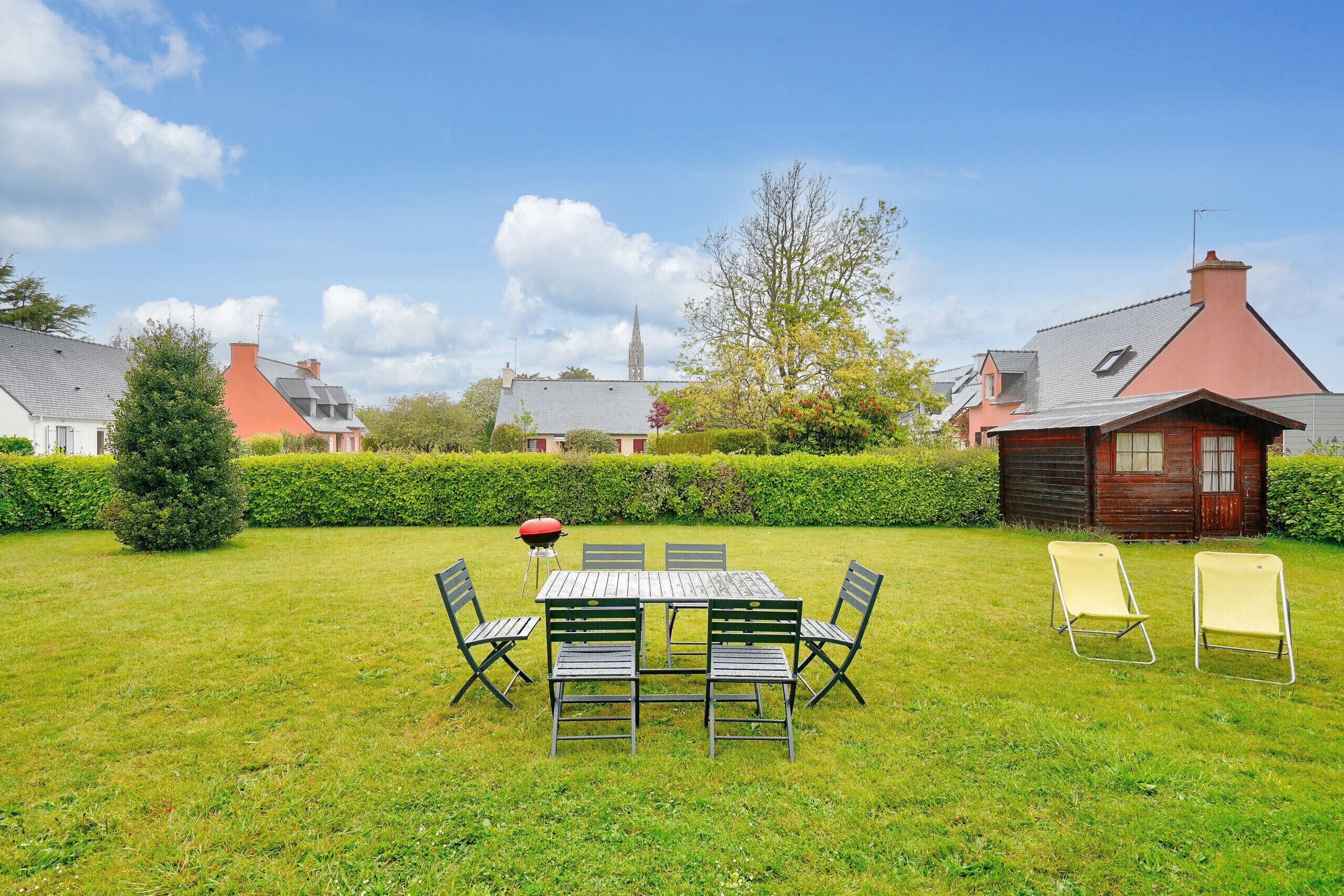 A grassy outdoor area with a wooden table and chairs, a barbecue grill, and a small wooden building.