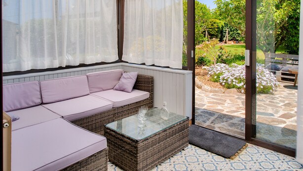 Un solarium con un área de estar acolchada, una mesa con superficie de vidrio y vista a un jardín a través de una puerta de cristal.
