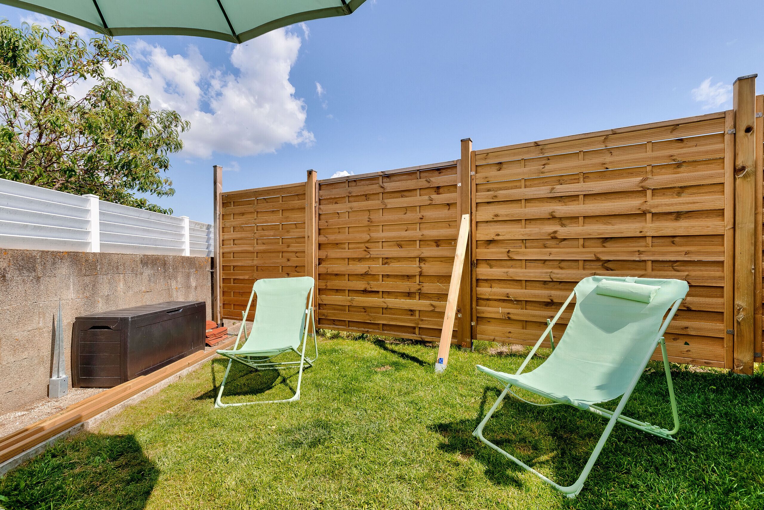 Patio trasero con valla de madera, dos sillas de jardín verdes y una parrilla.