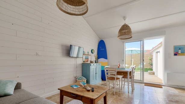 A living room with a sofa, coffee table, and a dining area with chairs and a table. There is a TV on the wall and a surfboard decoration.