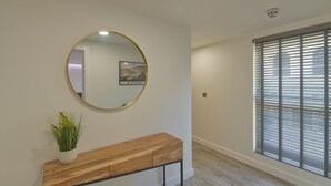 A hallway with a round mirror, a wooden console table with a plant, and a window with blinds. - Marina Walk, New Luxury Coastal Apartment in The English Riviera (Torquay)