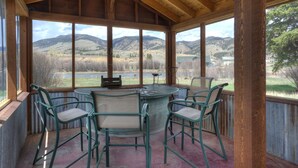 A covered patio with a table and chairs, large windows offering a view of a mountainous landscape. - Riverfront Access | Pet Friendly | Expansive Deck w/Mtn Views | ATV's Welcome (Creede)