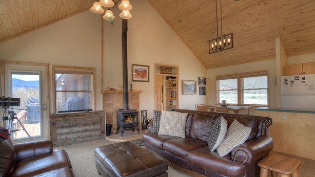 A cozy living room with a leather sofa, ottoman, and a fireplace. There is a wooden ceiling and a kitchen area visible in the background. - Riverfront Access | Pet Friendly | Expansive Deck w/Mtn Views | ATV's Welcome (Creede)