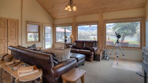 A cozy living room with leather sofas, a wooden ceiling, and large windows offering a view of the outdoors. - Riverfront Access | Pet Friendly | Expansive Deck w/Mtn Views | ATV's Welcome (Creede)
