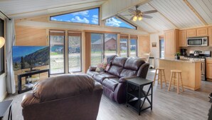 A spacious living room with a leather sofa, a coffee table, and a TV. The room has large windows offering a view of the outdoors.