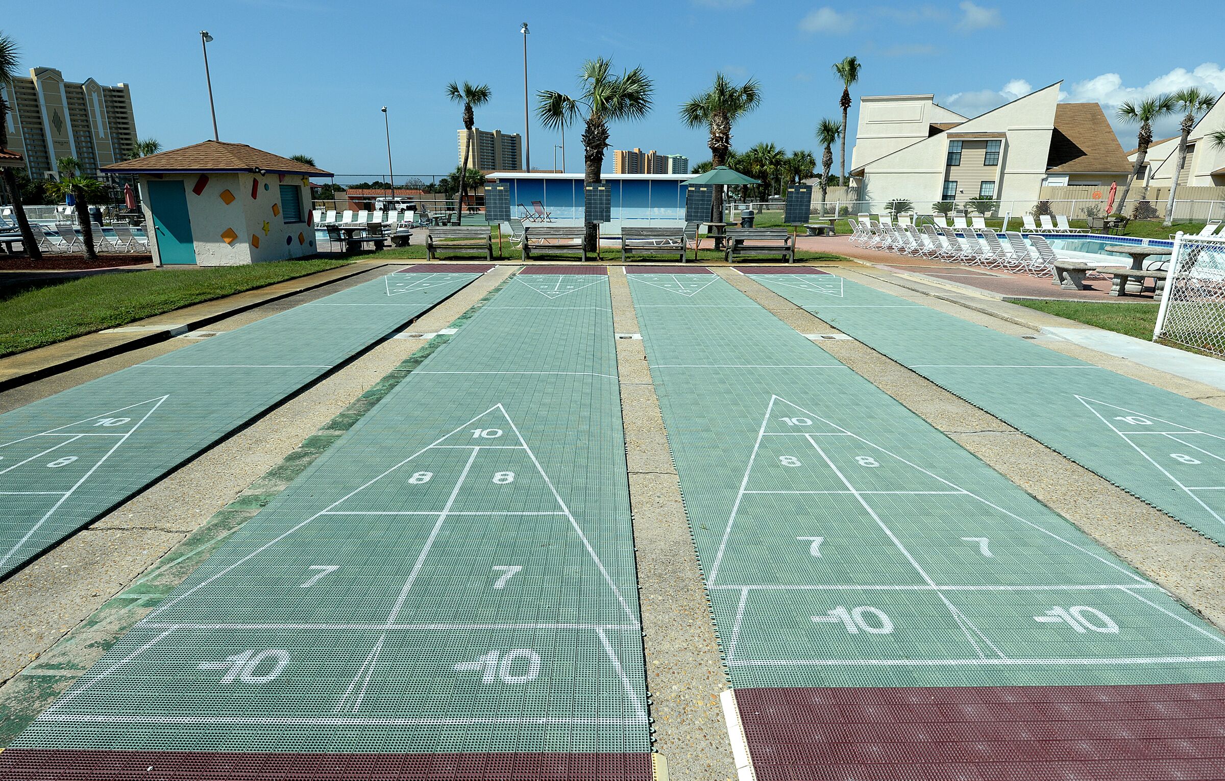 A shuffleboard court with green and red sections, surrounded by palm trees and a building with a blue roof.