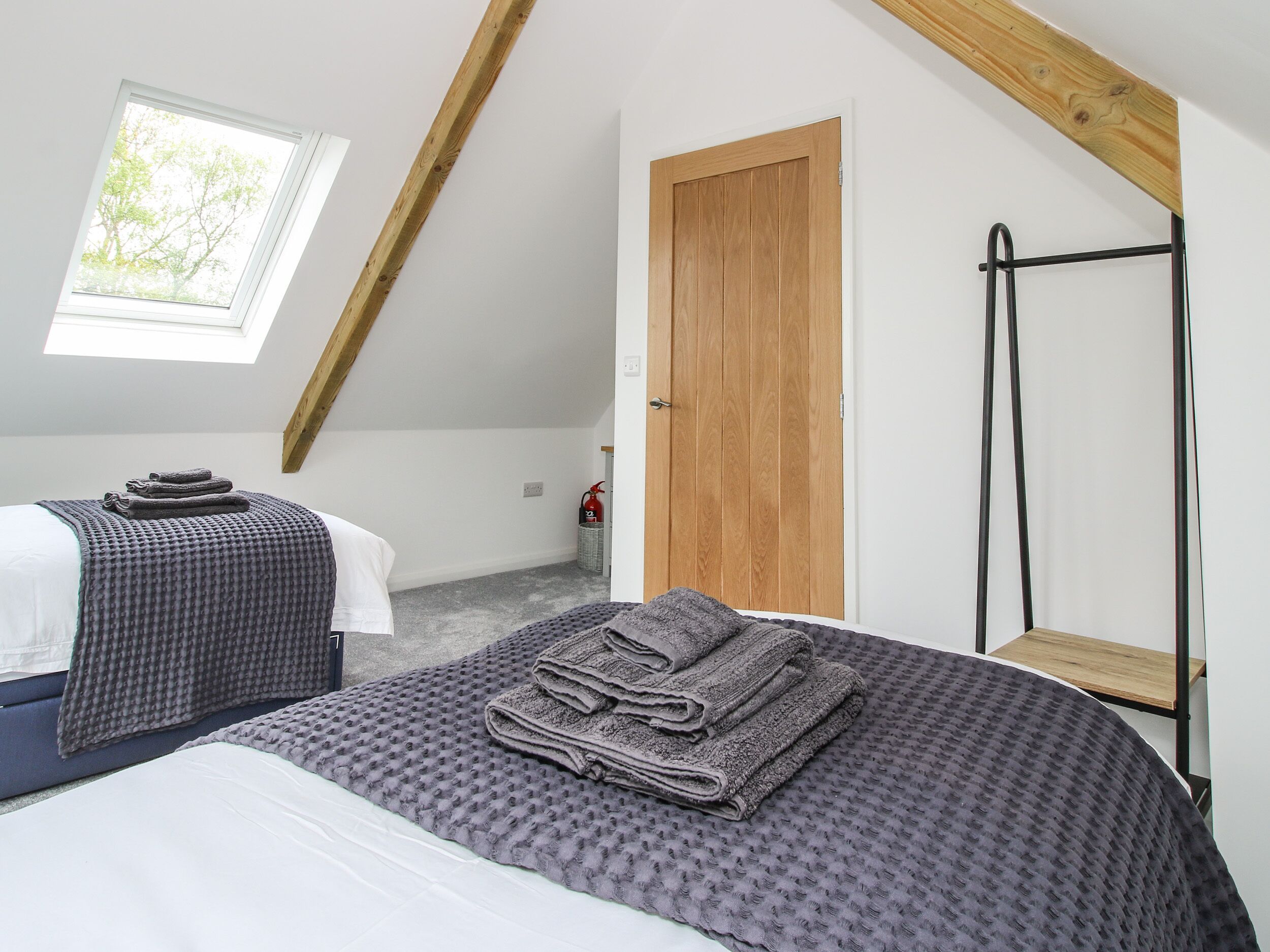Une chambre avec un plafond en pente, une porte en bois, un étagère en métal et une fenêtre donnant sur les arbres.