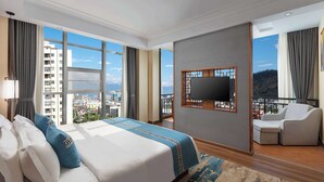 Une chambre d’hôtel dotée d’un grand lit, d’un téléviseur fixé au mur, d’un balcon offrant une vue sur la ville et d’un coin salon avec un fauteuil et un pouf.