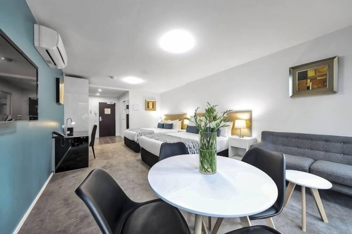 A hotel room with a bed, a dining table with chairs, a sofa, a small kitchen area, and a bathroom.
