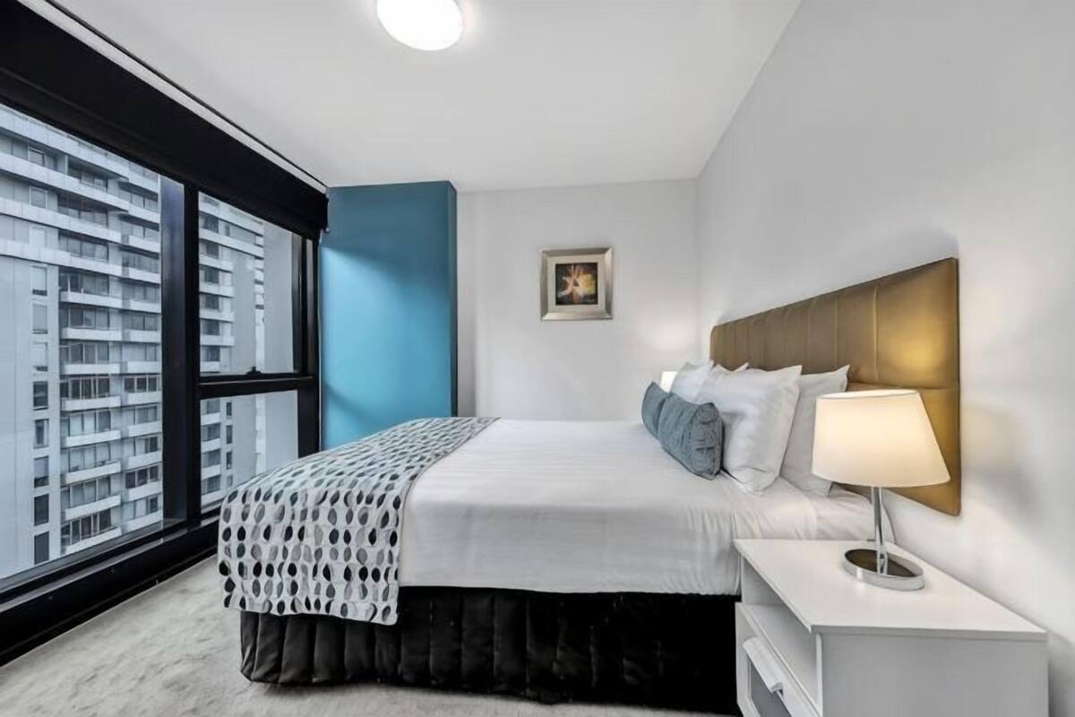 A modern hotel room with a large window, a bed with a headboard, a bedside table with a lamp, and a small picture on the wall.
