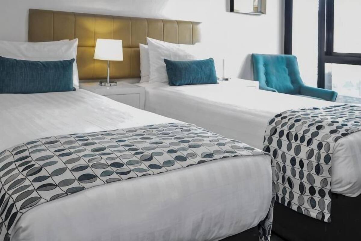 A hotel room with two beds, a nightstand with a lamp, a window with a view, and a blue chair.