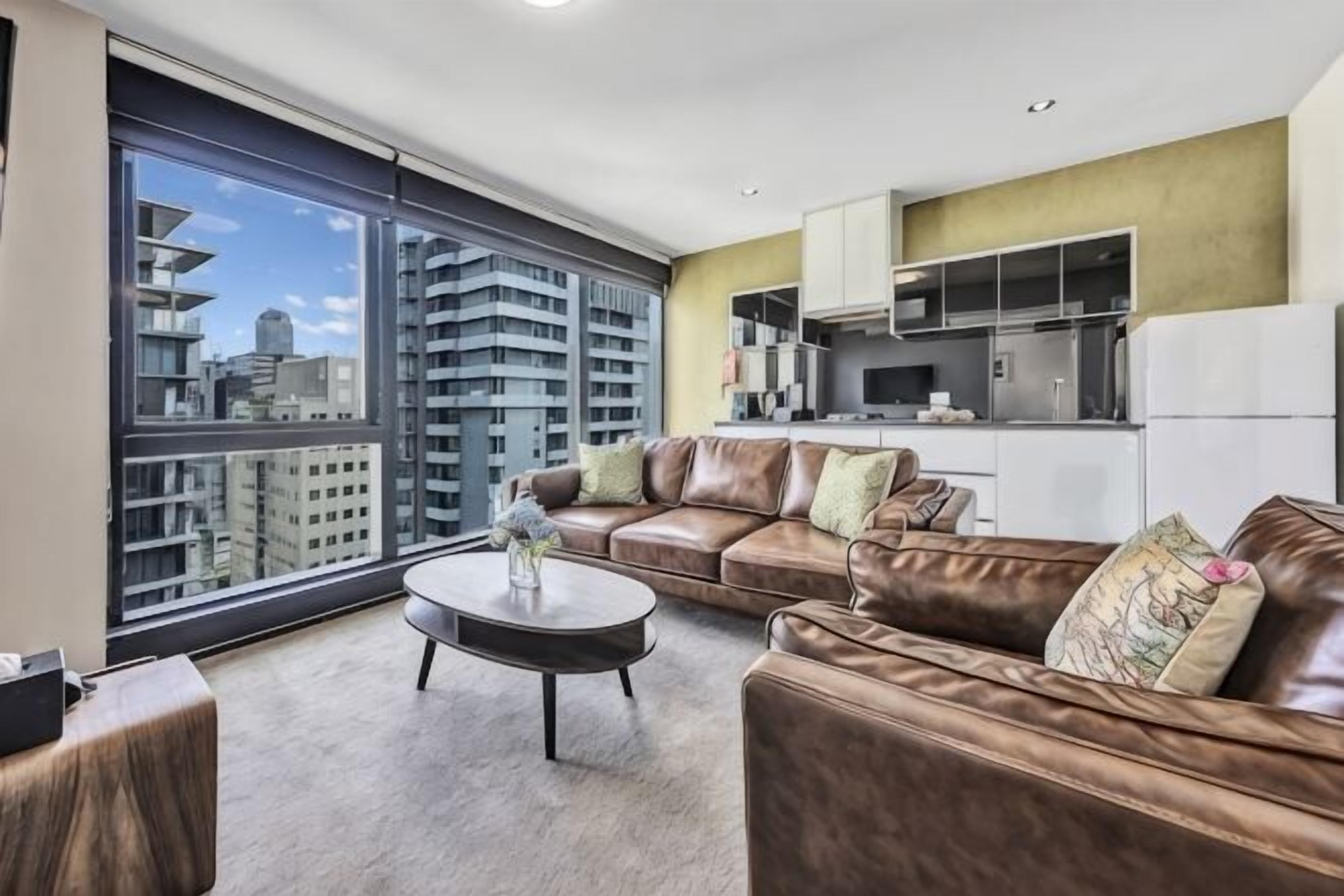 A modern living room with a leather sofa, a coffee table, and a large window offering a city view.