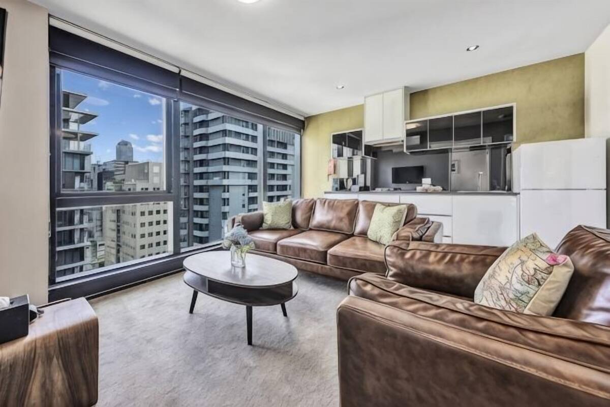 A modern living room with a leather sofa, a coffee table, and a large window offering a city view.
