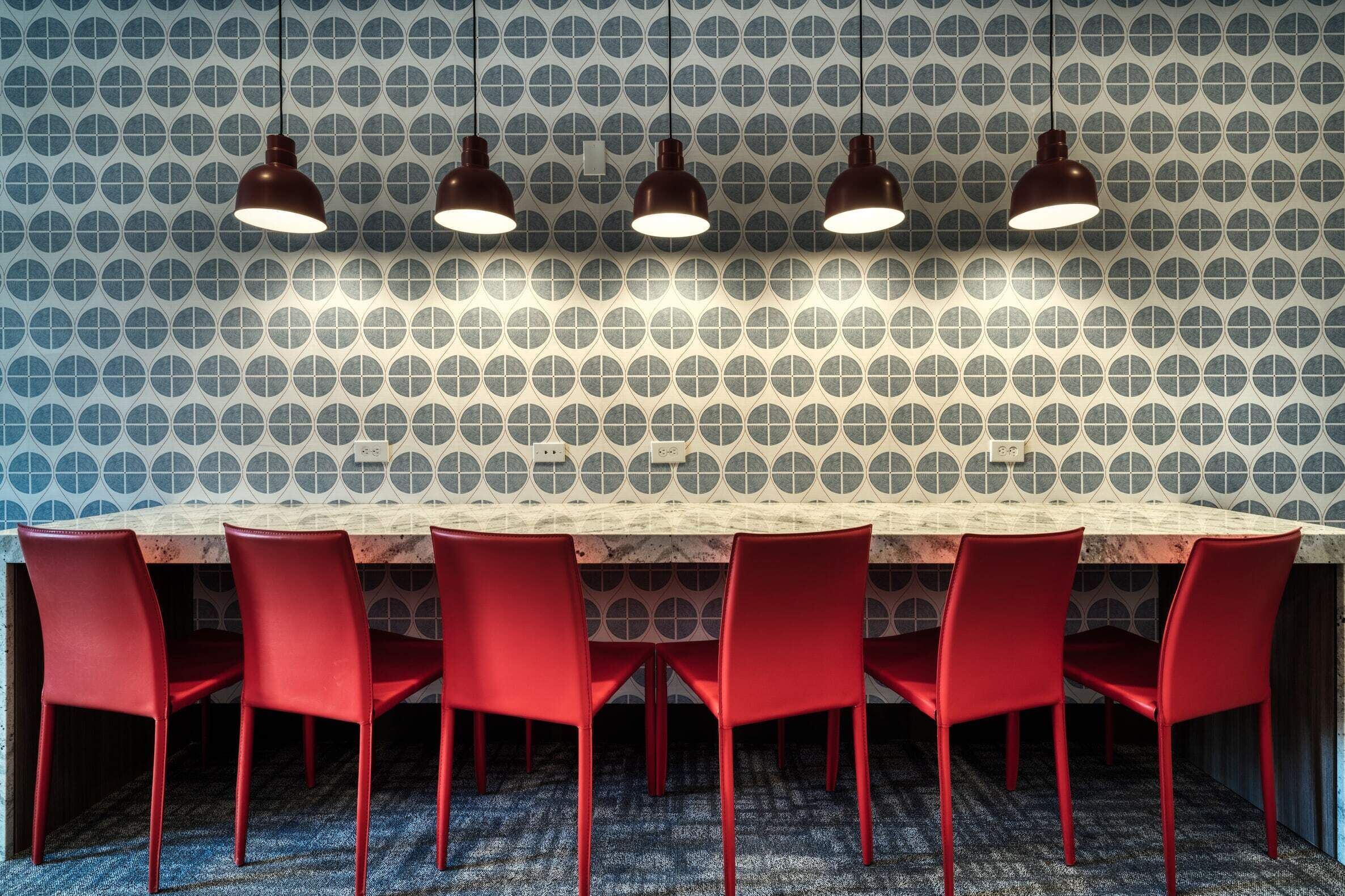 Een vergaderruimte met een lange tafel, zes rode stoelen en drie hanglampen.