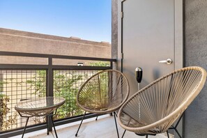 A balcony with two wicker chairs and a small table, a door with a handle, and a view of a building and greenery. - CozySuites at Westgate (Glendale)