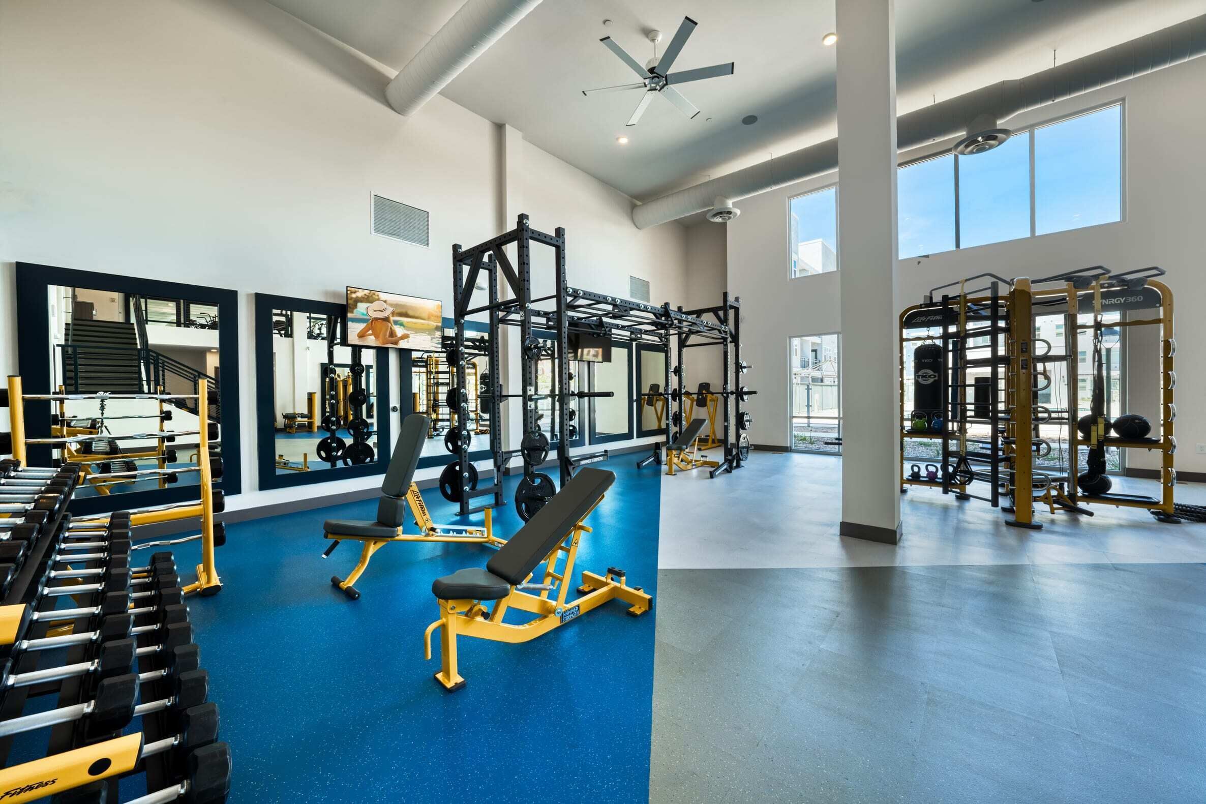 Et rummeligt fitnessrum med styrkeløftudstyr, spejle og store vinduer.