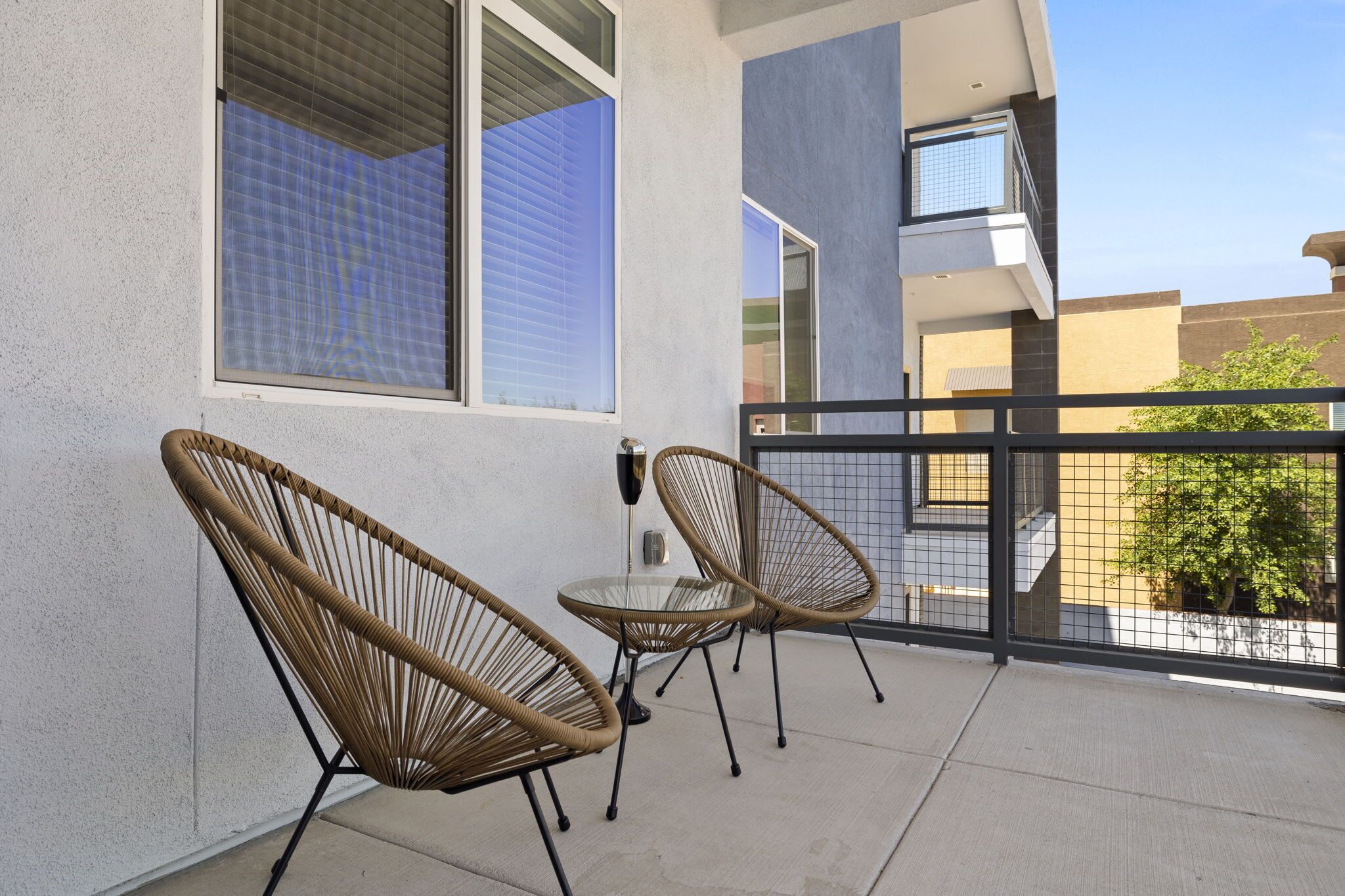 A balcony with two wicker chairs and a small table, a window with blinds, and a view of neighboring buildings.