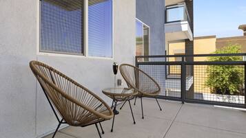 A balcony with two wicker chairs and a small table, a window with blinds, and a view of neighboring buildings.