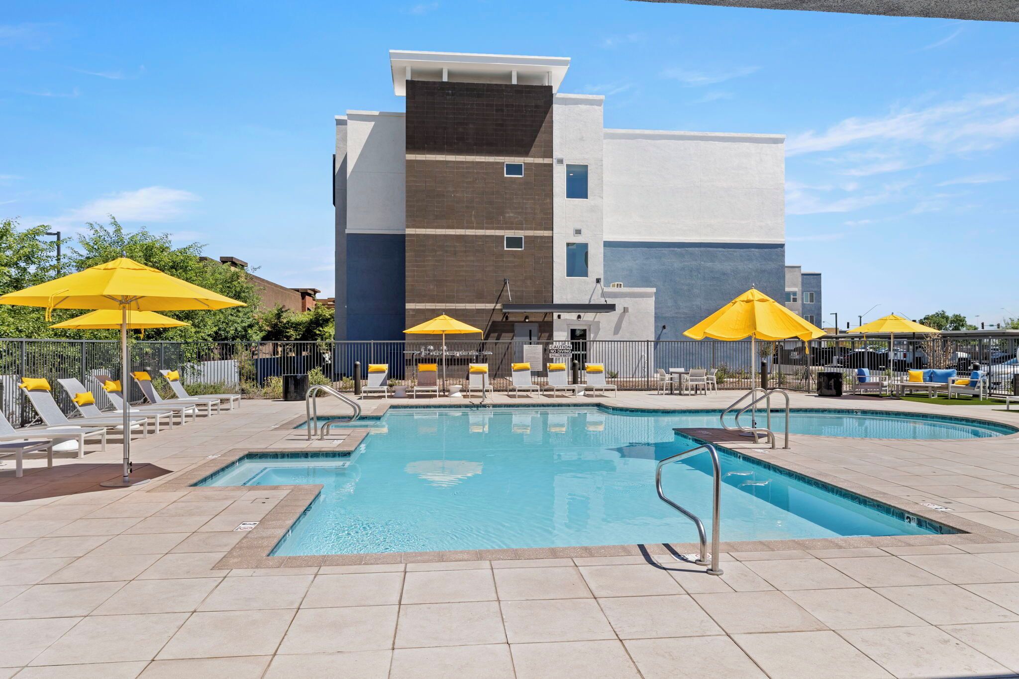 A swimming pool with sun loungers and umbrellas, surrounded by a modern building with a flat roof.